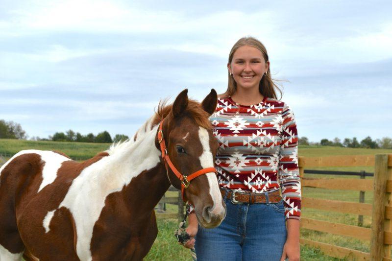 Ashtyn Dunn stands holding a horse's bridle on a farm